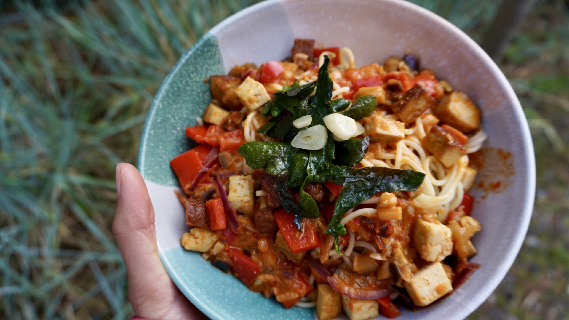 A deep plate of spaghetti topped with vibrant green pieces of bladderwrack algae and sautéed garlic. The pasta is coated in a rich, red ajvar sauce, interspersed with golden brown cubes of crispy tofu and soft, translucent pieces of eggplant. The dish is garnished with freshly ground black pepper, and the colorful medley of ingredients creates a visually appealing, nutritious meal.