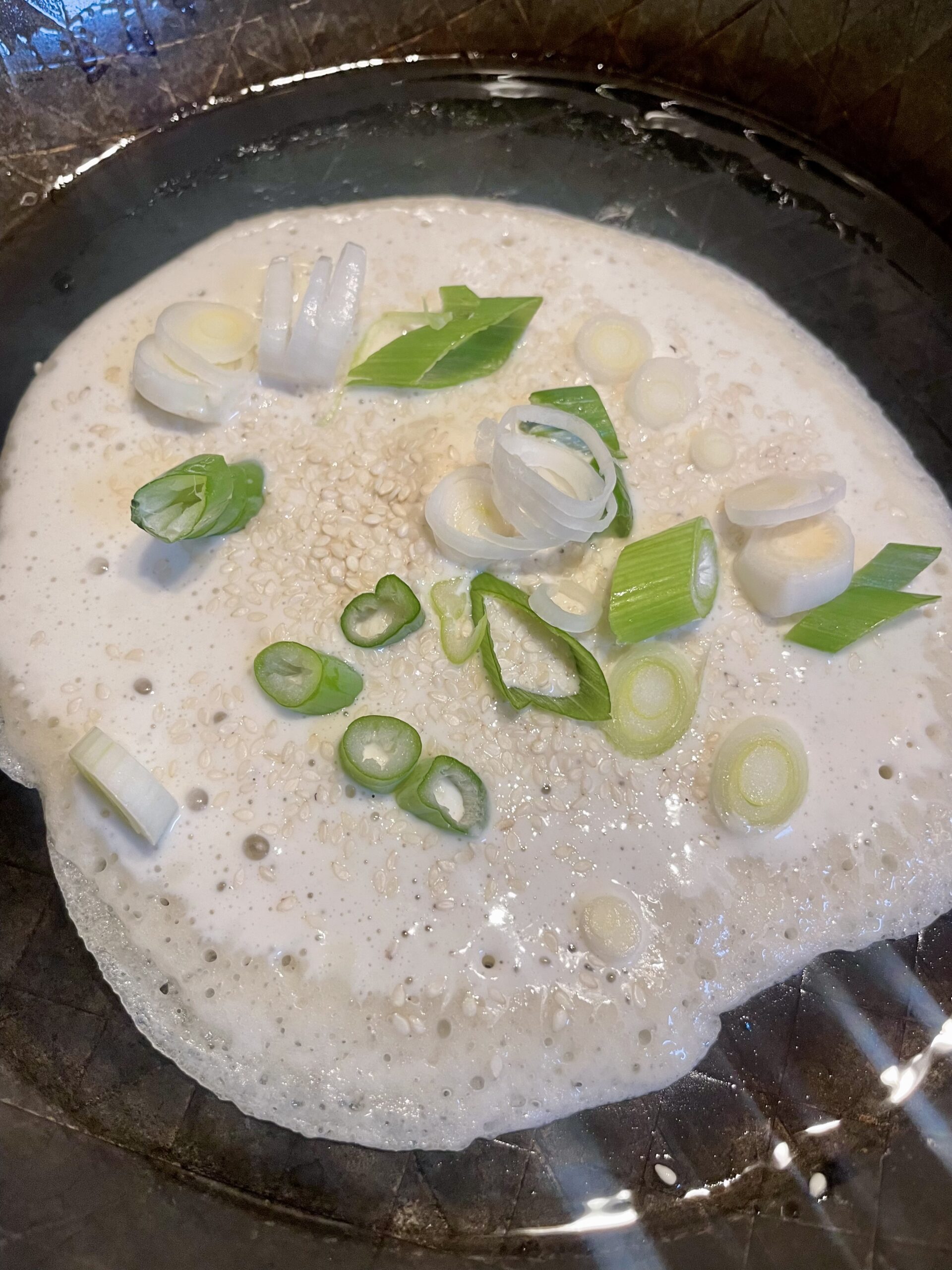 Pouring thin layer of sourdough discard into a heated skillet to create a crispy pancake surface. Sesame seeds being sprinkled evenly onto the uncooked batter for added flavor and texture.